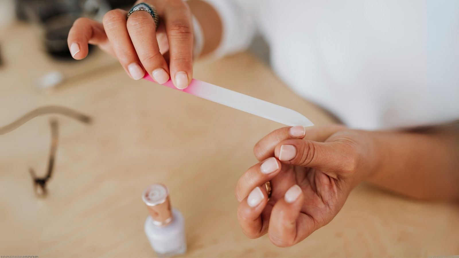 Close-up of a person using a glass nail file to shape natural nails, with nail polish bottle in the background